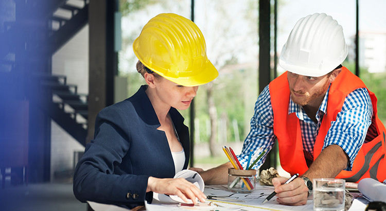 uma mulher de capacete amarelo e um homem de capacete branco conversando e analisando um projeto.